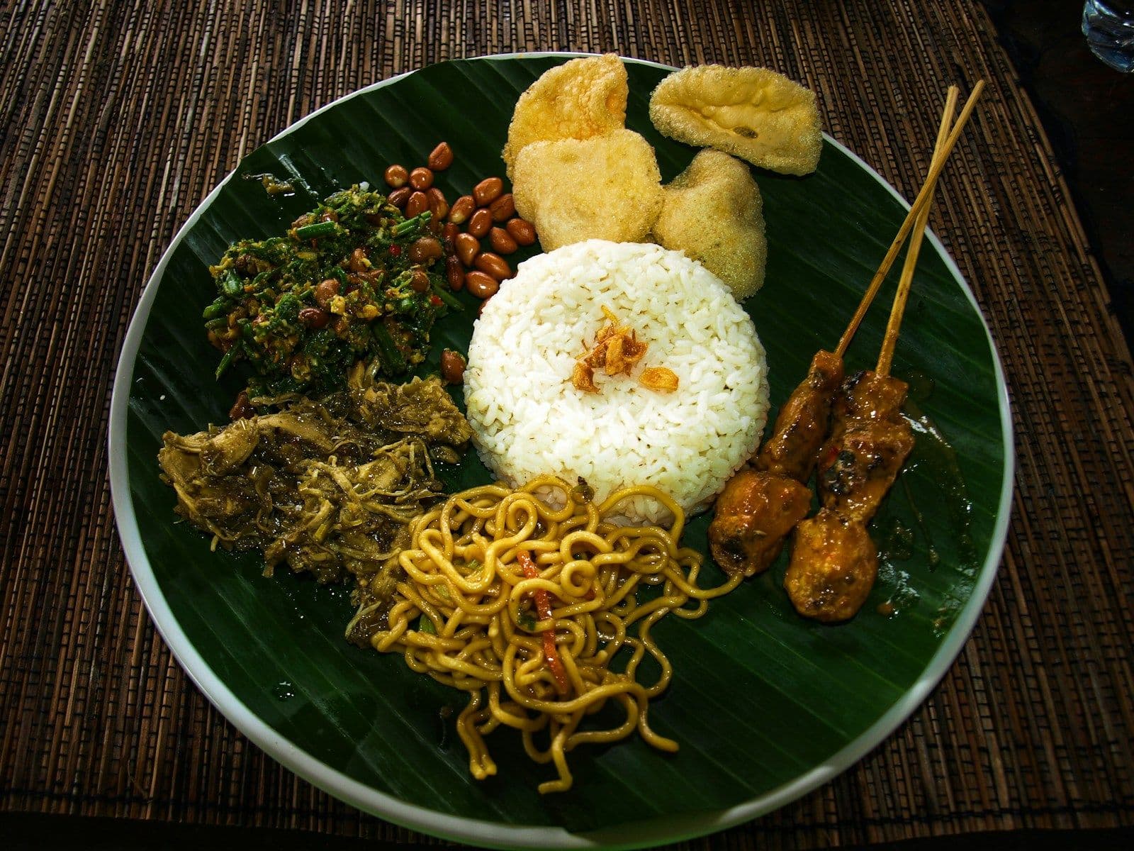 Sepiring nasi dengan sate, mi goreng, sayur, kacang, dan kerupuk beralas daun pisang