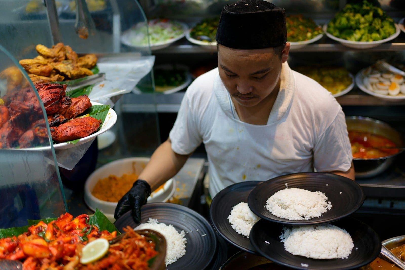 Pegawai rumah makan berpeci menyiapkan piring-piring nasi di depan etalase lauk